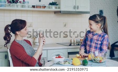 Young mother talking photo on smartphone camera of her cute daughter cooking cutting vegetables in the kitchen at home. Family, cook, and people concept