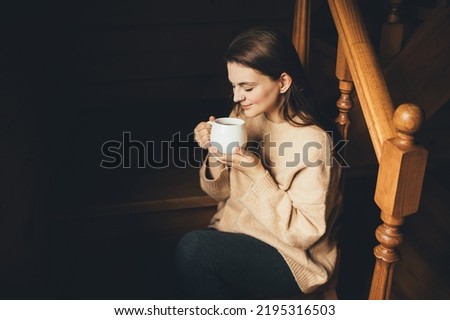 Young woman wearing warm knitted sweater drinking hot tea in a cozy wooden country house.