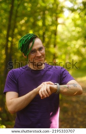 Man using smart watch in park. Male in violet t shirt with glasses and green deadlocks checking notifications on smart watch on summer day in park.