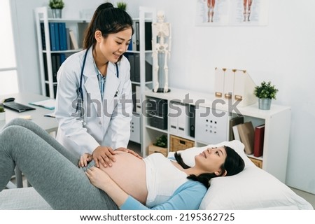 friendly Asian female doctor is giving a health checkup by touching the pregnant woman’s abdomen as she is lying on treatment couch in the hospital. Royalty-Free Stock Photo #2195362725