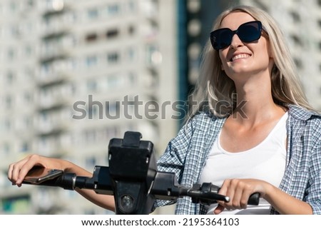 Portrait of a young happy blonde woman in a white T-shirt, shirt, sunglasses with a scooter in her hands on a sunny summer day in the city