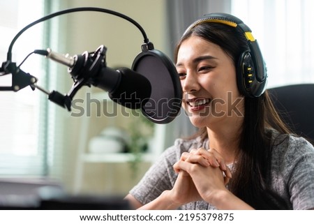Asian attractive audio DJ woman speaks into microphone to broadcasting. Young beautiful female blogger influencer wearing headphones and recording morning news podcast show for radio at home studio.
