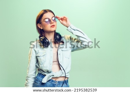 Portrait of young girl, student in casual clothes, sunglasses and headphones posing isolated over green background. Concept of youth, fashion, lifestyle, emotions, facial expression. Copy space for