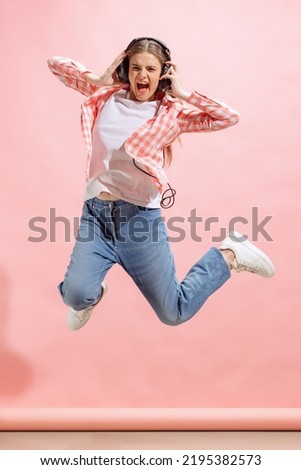 Portrait of excited young girl in checkered shirt listening to music in headphones and jumping isolated over pink studio background. Concept of youth, fashion, lifestyle, emotions, facial expression