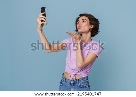 White woman sending air kiss while taking selfie photo at cellphone isolated over blue background