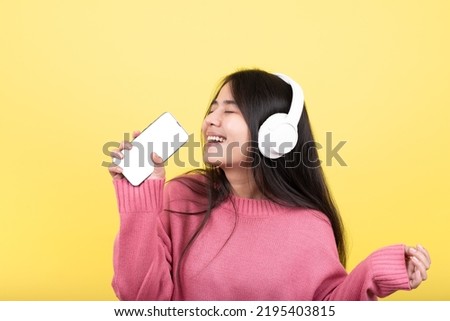 Happy Asian girl wearing headphones listening to relaxing mobile phone music with eyes closed on lonely yellow studio background. isolated