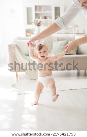 A charming baby girl in diapers takes her first steps at home by the hand with her mother, a child learns to walk at home in a bright living room, a healthy child at home Royalty-Free Stock Photo #2195421753