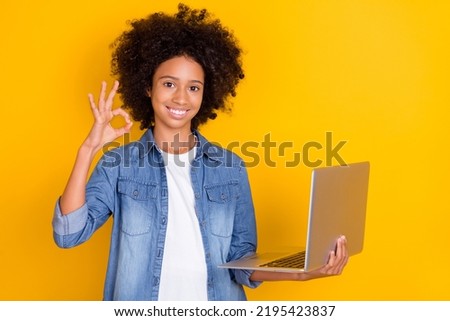 Portrait of beautiful trendy cheery teen girl using laptop showing ok-sign rate isolated over bright yellow color background
