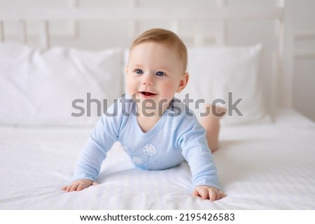 a cute healthy little baby is lying on a bed on white bedding at home in a blue bodysuit. The kid looks at the camera, smiles Royalty-Free Stock Photo #2195426583
