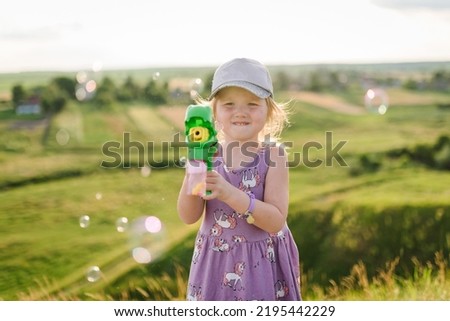 Happy girl blowing and catching soap bubbles in nature. Child walks on the green grass in the field at sunset. The kid is playing and smiling in the mountains.