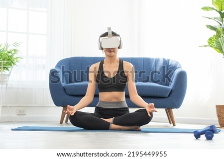 Asian woman using VR virtual reality glasses headset greeting friends in yoga class.Young female goggle using VR enjoy exciting exercise yoga watching 360 degree at home.Virtual Reality Metaverse