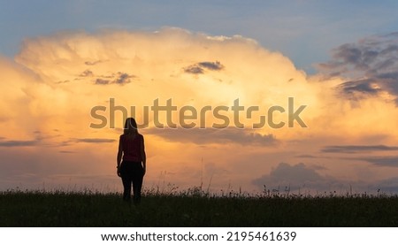 Silhouette of young caucasian woman in shirt walking through grass against sunset cloud sky