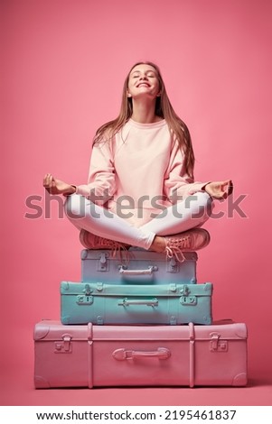 Keep calm and have a vacation. Traveling concept. Young pretty woman meditating in lotus pose on the luggage valises. Pink background.