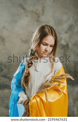 Little girl in ethnic embroidered dress covered with Ukrainian flag holding ears of wheat against dark loft background.