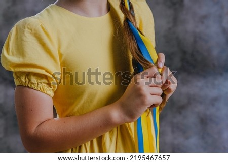 Close up of little girl braiding her hair with yellow blue ribbon against dark loft background with blank space for text.