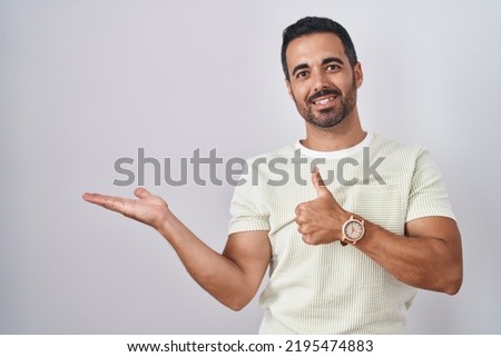Hispanic man with beard standing over isolated background showing palm hand and doing ok gesture with thumbs up, smiling happy and cheerful 