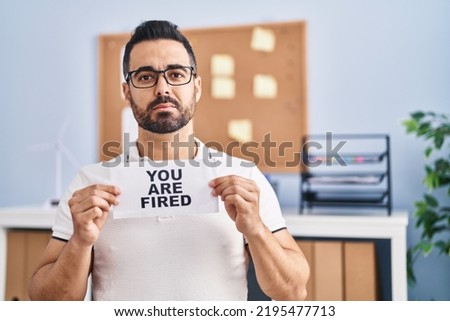 Young hispanic man with beard holding you are fired banner at the office relaxed with serious expression on face. simple and natural looking at the camera. 