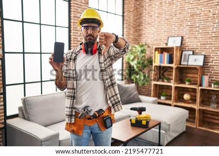 Young hispanic man with beard working at home renovation holding smartphone with angry face, negative sign showing dislike with thumbs down, rejection concept 
