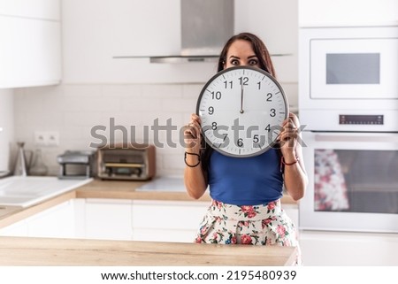 Woman stands in the kitchen holding big clock showing exactly twelve o'clock.