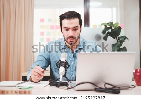 Young man host sitting at table, streaming audio podcast using microphone and laptop in studio,  Social media, podcasting, blogging concept. Cheerful casual caucasian employee.