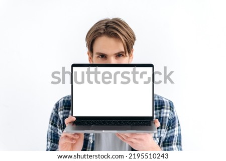Amazed excited caucasian young man peeking out from behind laptop, looks surprised at camera, stands on isolated white background, holds an open laptop with blank white screen, copy space, mock-up Royalty-Free Stock Photo #2195510243