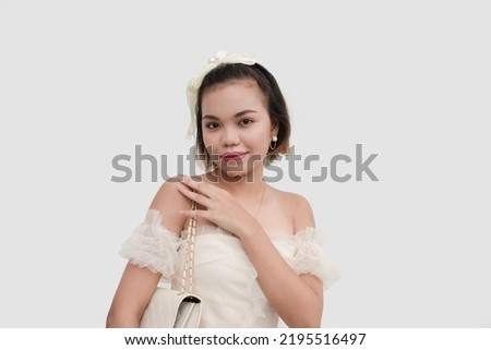 A timid and petite woman wearing a white off shoulder lace and a bow on her head holding the strap of her white bag isolated on a white background.