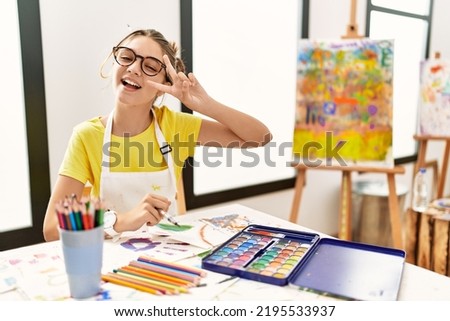 Young brunette teenager at art studio doing peace symbol with fingers over face, smiling cheerful showing victory 