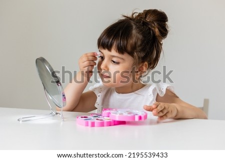 cute little girl in a white T-shirt is sitting at the dressing table in front of the mirror and applying makeup. like Mom. girl playing with makeup. space for text. High quality photo