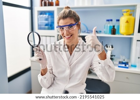 Young woman working at scientist laboratory holding magnifying glass smiling happy and positive, thumb up doing excellent and approval sign 