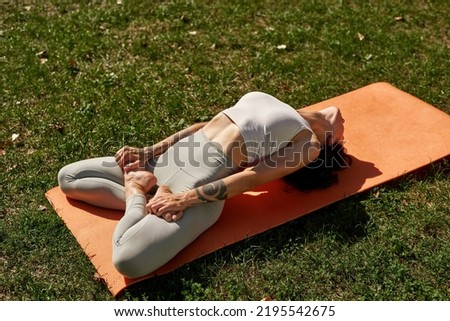 Young sportswoman practicing yoga in fish pose in sunny park. Attractive slim girl with tattoos wearing sportswear sitting on fitness mat on green lawn. Concept of healthy lifestyle