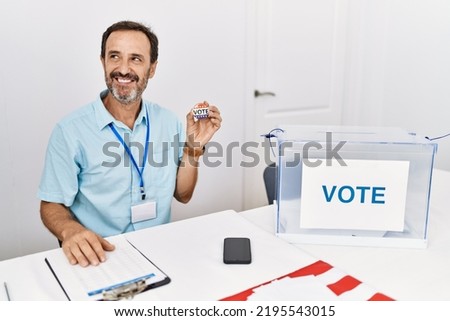 Middle age man with beard sitting by ballot holding i vote badge smiling looking to the side and staring away thinking. 