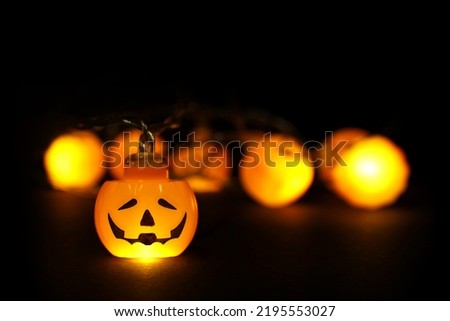Garland lights of glowing halloween orange pumpkins with copy space above and below on black background. Blurred bokeh lights, one pumpkin face focused, close up. Festive holidays Halloween concept. Royalty-Free Stock Photo #2195553027