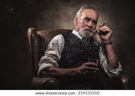 In chair sitting senior business man with cigar and whisky. Gray hair and beard wearing blue striped gilet and tie. Against brown wall. Royalty-Free Stock Photo #219555310