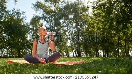 European elderly woman drinking water and resting after sports training in green sunny park. Sportive female with closed eyes wearing sportswear sitting on fitness mat on lawn. Healthy lifestyle