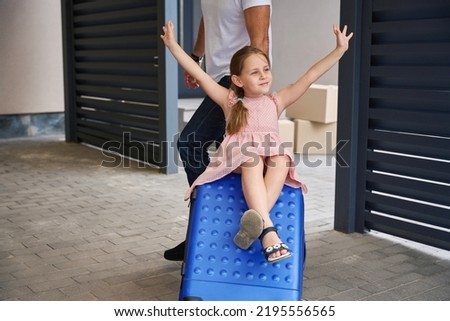 Father rolls happy daughter on blue suitcase near the house