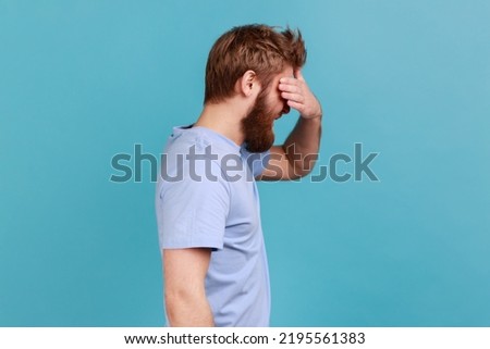 Don't want to look at this. Side view of bearded man covering eyes and turning with disgust from something shameful, scared afraid to see. Indoor studio shot isolated on blue background. Royalty-Free Stock Photo #2195561383