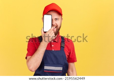 Portrait of happy smiling joyful bearded worker man standing and covering eye with cell phone with blank screen for promotional text. Indoor studio shot isolated on yellow background.
