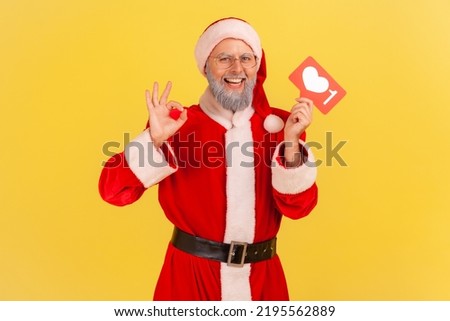 Portrait of smiling elderly man with gray beard wearing santa claus costume recommending to follow interesting blog, showing okay sign. Indoor studio shot isolated on yellow background.