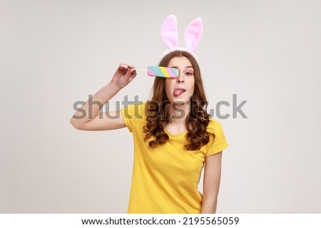 Portrait of adorable funny teenager in yellow casual T-shirt with pink bunny ears looking at camera and covering eye with ice cream. Indoor studio shot isolated on gray background.