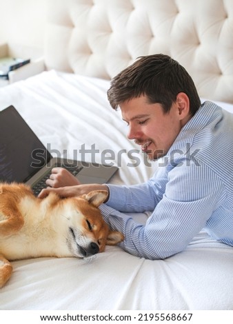 Young European developer male is working in a home office outsource. Online card payment. Emotional support Japanese breed shiba inu dog in bed. Laptop working vertical copy space Royalty-Free Stock Photo #2195568667