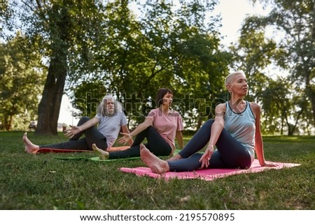 Three focused elderly caucasian friends practicing yoga in sage marichi's pose on fitness mats on meadow. Sportive man and women barefoot. Friendship. Healthy lifestyle. Sunny green park