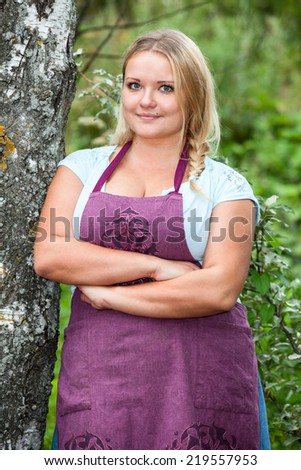 Smiling Caucasian woman in apron with clasped hands leaning birch, outdoors