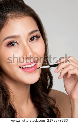 Close up young ardent woman with healthy fair skin applying lipstick on her lip while looking at camera. Beauty concept with cosmetic and makeup.