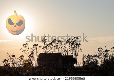 Halloween concept. Mysterious abandoned house at sunset in dark light. transparent Pumpkin Jack lantern instead of the moon. Weeds are in the foreground. Terror and fear concept. Selective focus.