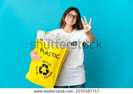 Woman holding a bag full of bottles to recycle isolated on blue background happy and counting three with fingers