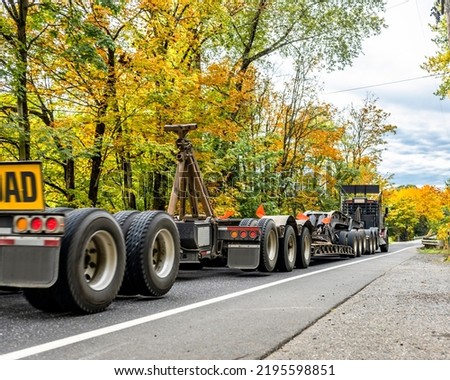 Industrial big rig hauler semi truck with oversize load sign transporting oversized commercial cargo on two long step down semi trailers running on the road with autumn forest in Columbia Gorge
