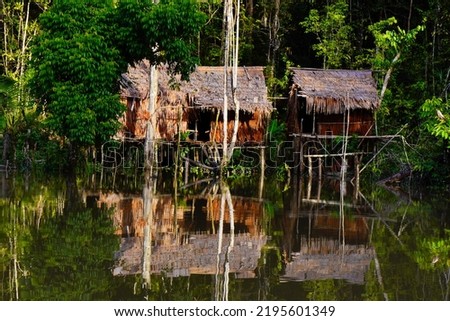 Three cottages with natural materials located on the banks of a river with a dense tropical forest background give the impression of magic, naturalness, peace and silence Royalty-Free Stock Photo #2195601349