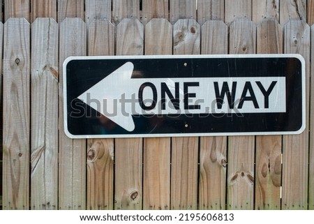 A metal directional traffic sign is attached to an unfinished wooden fence. The black and white arrow has the words one way in capital letters with black text. The pointing arrow is on an urban road. 