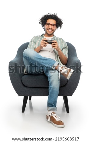 technology, people and leisure concept - happy smiling young man in glasses with gamepad sitting in chair and playing video game over white background