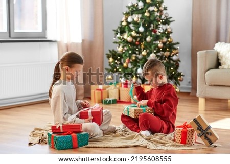 christmas, winter holidays and childhood concept - happy girl and boy in pajamas opening gifts sitting on floor in front of each other at home